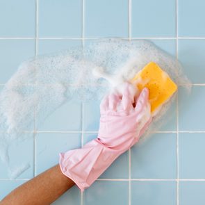 Gloved hand scrubs blue tiled wall with a yellow sponge, creating soapy foam.