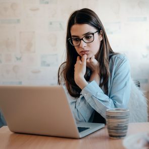 A woman sits focused on a laptop in a cozy room, with a striped mug and patterned wallpaper in the background.