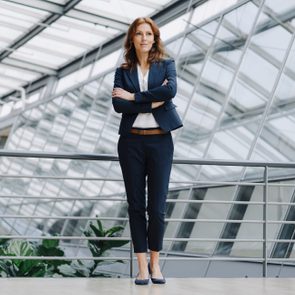 A woman stands confidently with crossed arms, wearing a suit, in a modern glass-roofed building with metal railings and plants.
