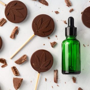 Chocolate lollipops with leaf imprint are scattered on a surface, surrounded by chocolate chunks, next to a green glass dropper bottle.