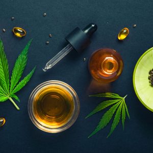 Cannabis leaves lie next to a dropper bottle and oils on a dark background, seeds in green bowl nearby.
