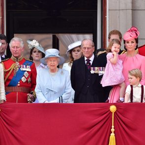 Group stands on a balcony, some in uniforms and formal attire, surrounded by decorative red drapery in a ceremonial setting.