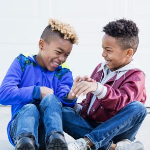 Two children are laughing and sitting on the ground, wearing colorful jackets and jeans, in a bright outdoor setting.