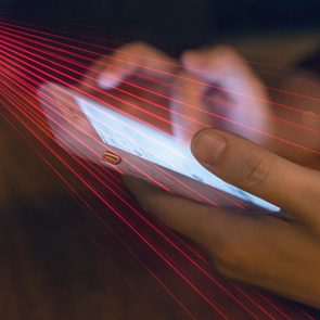 Hands hold a phone, emitting a bright screen, with red laser lines crossing diagonally, in a dimly lit setting.