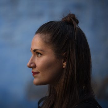 Woman gazes forward, side profile, with dark hair in a top knot, against a blurred blue background.