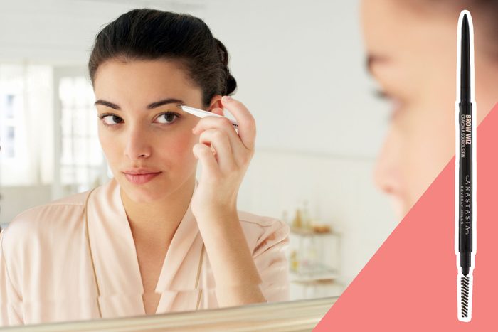 A woman uses an eyebrow pencil, looking into a mirror in a bright room. Text reads 