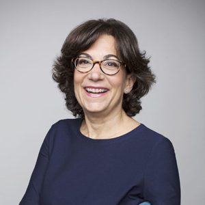A person smiles while sitting against a neutral background, wearing glasses and a dark blue top.