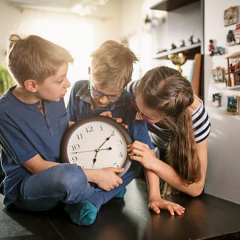 Clock held by three children, gathered and examining it closely, inside a cozy room with decorative wall items and soft, natural lighting.
