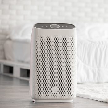 Air purifier sits on a wooden floor, operating quietly. Nearby is a white bed with a brick-patterned wall in a cozy bedroom.