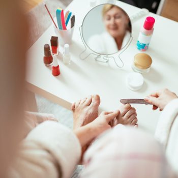 Hands file toenails, surrounded by nail polish bottles and a mirror reflecting a blurred face, on a small white table.
