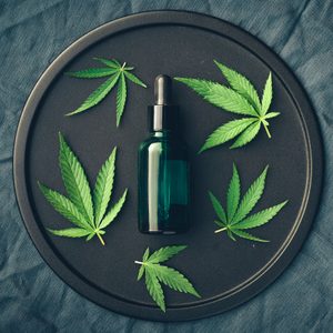 Green dropper bottle sits on a dark tray, surrounded by cannabis leaves, placed on a blue fabric background.