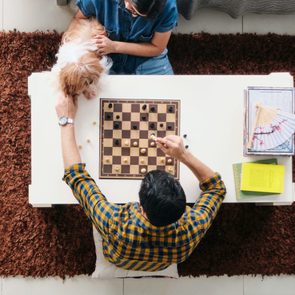 Chessboard sits on a table, played by two people; one person observes while petting a dog. A book and fan are nearby.