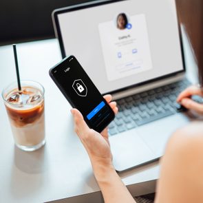 A smartphone shows a login screen with a lock icon while a person uses a laptop beside an iced coffee on a table.