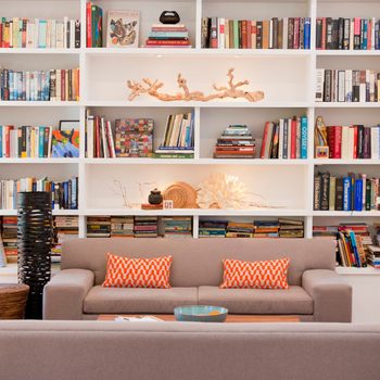 Sofa sits facing bookshelves filled with books; accented by orange pillows and decorative elements. Soft light illuminates the cozy space.