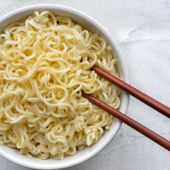 A bowl of cooked noodles sits with wooden chopsticks resting on it, placed on a light-colored surface.