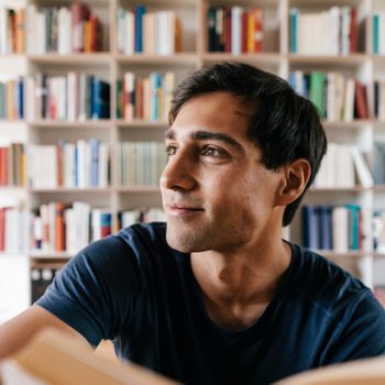 Person reading, smiling, surrounded by a colorful bookshelf in a bright room.