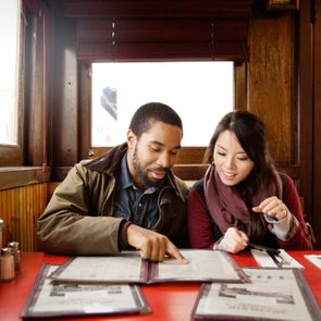 Two people review menus at a red table in a cozy wooden restaurant booth. Ketchup and utensils are placed nearby.