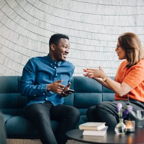 Two people sit talking on a blue couch, one holding a phone, with books and a candle on a table in front of them.