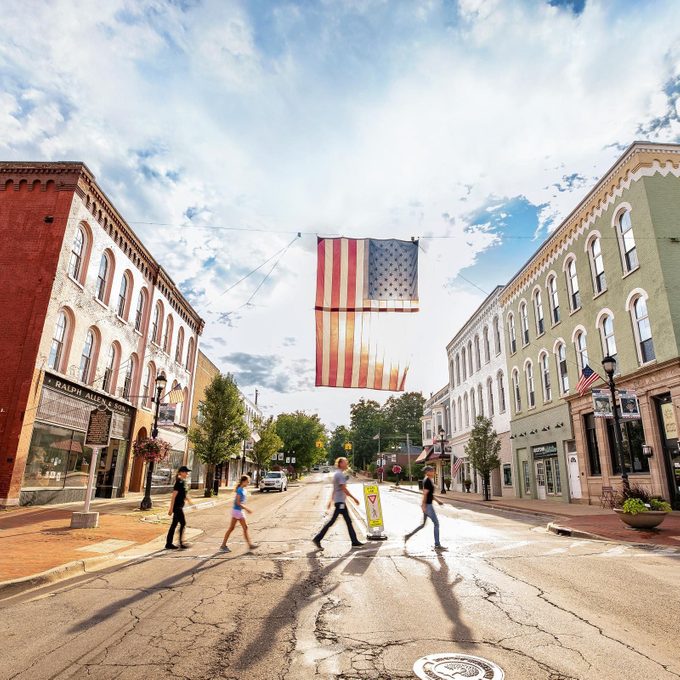 Flag hangs above pedestrians crossing a small town street, bordered by historic buildings under a partly cloudy sky.