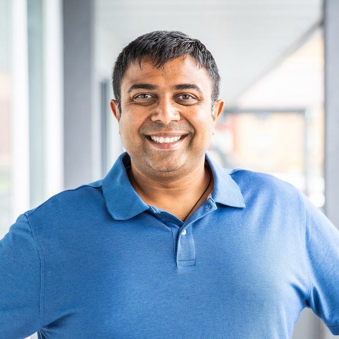 Smiling man stands confidently in a blue polo shirt, positioned in a brightly lit hallway with visible windows.
