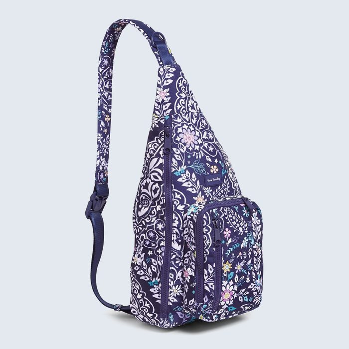 A floral-patterned sling bag stands upright, displaying multiple zippered compartments, on a plain light blue background. The text reads 