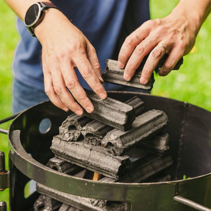 Hands arrange charcoal briquettes inside a round barbecue grill, set outdoors on green grass.