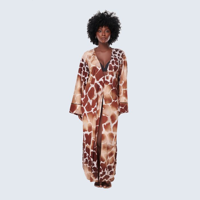 Woman stands wearing giraffe-patterned robe, revealing a dark top underneath, in front of a plain, light blue background.