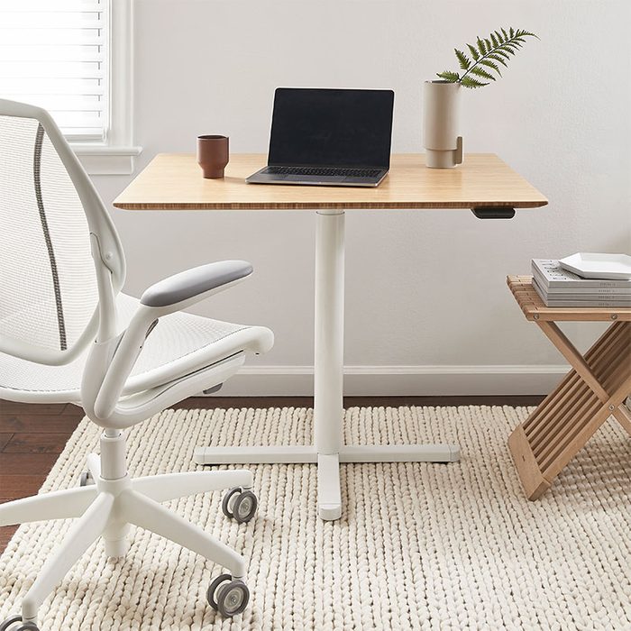 Laptop rests idle on a wooden desk, surrounded by a white chair, plant, and books, in a bright, minimalist room with a rug.