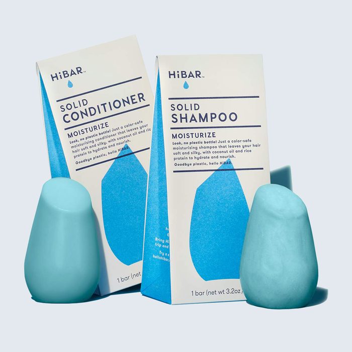Solid shampoo and conditioner bars stand upright beside packaging on a light blue background. Packaging text emphasizes moisturization and is plastic-free.