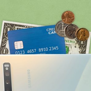 A smartphone conceals a credit card, dollar bill, and coins, resting on a green surface. The card number is 0123 4657 8910 2345.
