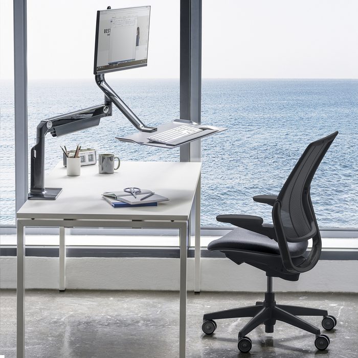 Standing desk setup with monitor and keyboard mounted, overlooking an ocean view through large windows. Office chair and desk accessories are present.
