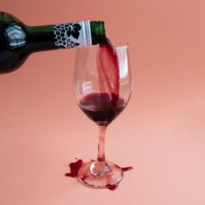 Wine bottle pouring into glass, overflowing onto table. Peach background.