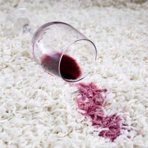 A wine glass lies on a carpet, spilling red wine and creating a noticeable stain on the white fabric.