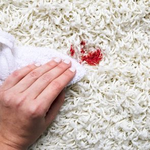 A hand uses a white cloth to dab a red stain on a cream-colored carpet.