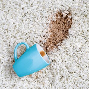 A blue mug lies on its side, spilling brown liquid onto a white, fluffy carpet, creating a noticeable stain on the fabric.