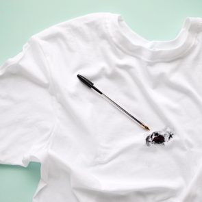 A white t-shirt lies flat with a black ink pen leaking onto it, creating a stain. The background is light green.