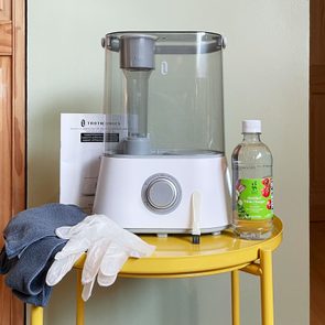 Humidifier sits idle on a yellow table, surrounded by cleaning gloves, cloth, manual, and a bottle of distilled white vinegar in a room setting.