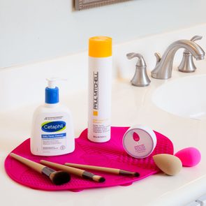 Toiletries and makeup brushes rest on a pink mat by a bathroom sink. Items include Cetaphil cleanser, Paul Mitchell shampoo, and beauty sponges.