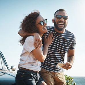 Two people laugh together, standing by a car near the ocean under a clear, sunny sky.