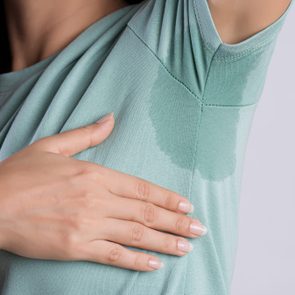 Hand touches underarm sweat stain on a teal shirt in a neutral background.