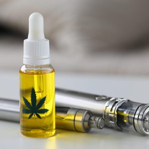 A glass dropper bottle with cannabis leaf design stands upright next to metallic vape pens on a white surface.