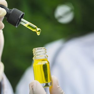 A dropper releases yellow liquid into a small glass bottle, held by gloved hands, against a blurred green background.