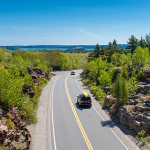 A car drives along a winding road through a lush, rocky landscape, with trees and distant water visible under a clear blue sky.