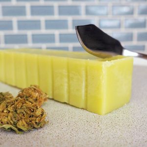 A block of green butter being sliced, next to dried herbs on a countertop, in front of a tiled wall.