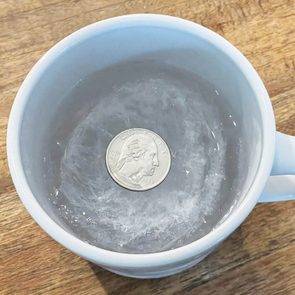 A coin rests on ice within a white mug, placed on a wooden surface.