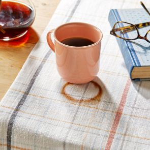 A pink mug holding coffee rests on a plaid cloth beside a coffee pot, book, and glasses on a wooden table.