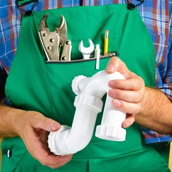 Hands holding a white plumbing pipe, while wearing a green apron with tools in the pocket, against a plaid shirt background.