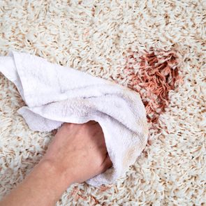 Hand cleans white carpet with a cloth, removing a reddish stain in a close-up view.