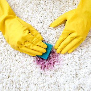 Gloved hands scrub a red stain with a sponge on a white carpet.