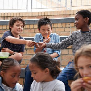 Children sit on steps, sharing snacks and interacting, smiling in a casual outdoor brick-walled area.
